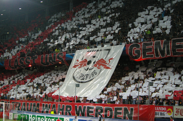 Ein Fußballstadion voller Zuschauer, einige sitzen und einige stehen, mit Bannern, einem Torpfosten und Scheinwerfern.