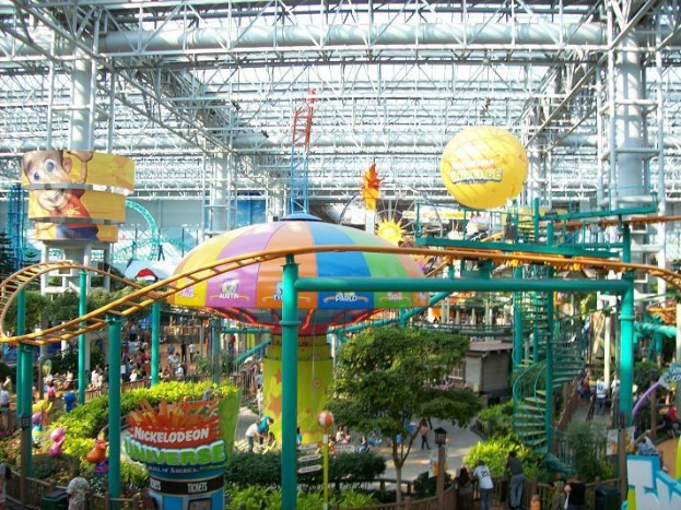 Ein großer Indoor-Wasserpark mit einer Achterbahn, umgeben von Menschen, Bäumen, Pflanzen und einer einladenden Decke mit Stangen und Lichtern.