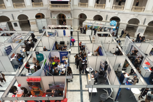 Große Besuchergruppe auf einer internationalen Fachmesse mit Ständen, Bannern und Luftballons in einem Raum mit architektonischen Bögen und Säulen.