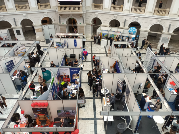 Große Besuchergruppe auf einer internationalen Fachmesse mit Ständen, Bannern und Luftballons in einem Raum mit architektonischen Bögen und Säulen.