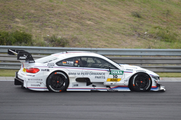 BMW M4 DTM-Rennwagen auf einer Rennstrecke mit Sponsor-Text, umgeben von grünem Gras und einer Absperrung im Hintergrund.