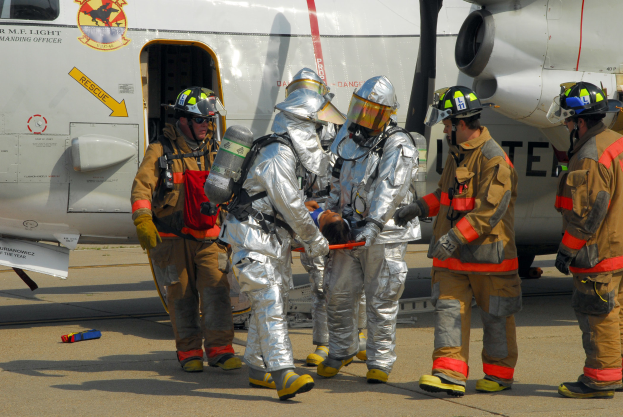 Eine Gruppe von Feuerwehrleuten in Helmen und Schutzausrüstung steht vor einem Flugzeug auf der Rollbahn, wobei einer ein Objekt hält und verschiedene Gegenstände auf dem Boden verstreut sind.