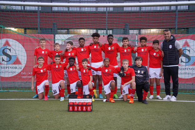 Eine Gruppe junger Männer in roten T-Shirts und schwarzen Shorts steht auf einem Fußballfeld vor einer Kiste roter Gegenstände, im Hintergrund sind Banner mit der Aufschrift "FC Rot Schwarz Eglaute - Champions League" zu sehen.