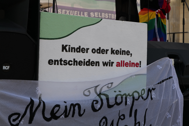Eine Person steht auf einer Bühne mit einem Schild, auf dem "Kinder oder keine, entscheiden wir allein" steht, einem Banner, einem Lautsprecher, einem Mikrofonständer und einem drapierten Tuch, mit einem Gebäude im Hintergrund.