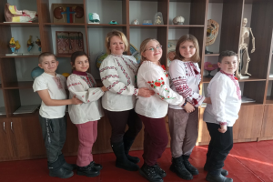 Eine Gruppe von Kindern in traditioneller ukrainischer Kleidung vor einem Regal mit Spielzeug, einem Skelett und anderen Gegenständen, was auf eine ukrainische Schulumgebung hinweist.