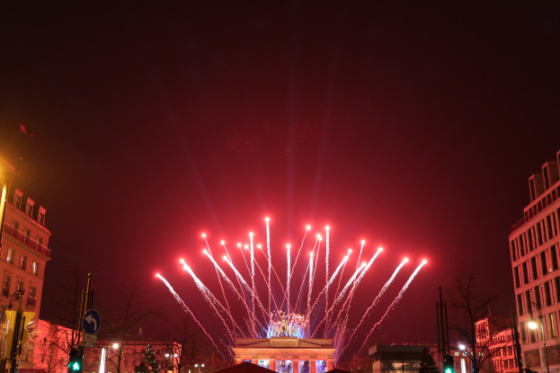 Das Bild zeigt eine nächtliche Straßenansicht in Berlin mit dem Brandenburger Tor in der Mitte, umgeben von beleuchteten Gebäuden, Bäumen, Verkehrsampeln, Schildern und Fahnen, während Feuerwerk den Himmel über dem Tor erhellt.