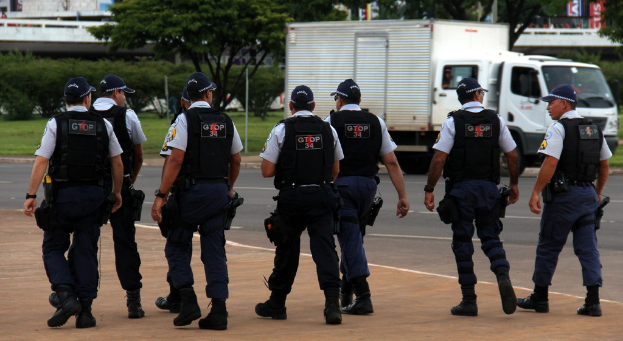 Eine Gruppe von Beamten in Uniform mit Mützen, Hemden, Schutzwesten, Uhren, Hosen und Schuhen, die auf einer Straße mit einem rechts geparkten Lkw neben einem Zaun, Gebäuden, Bäumen, Pflanzen, Polen und Bannern im Hintergrund und einer Brücke oben rechts gehen.