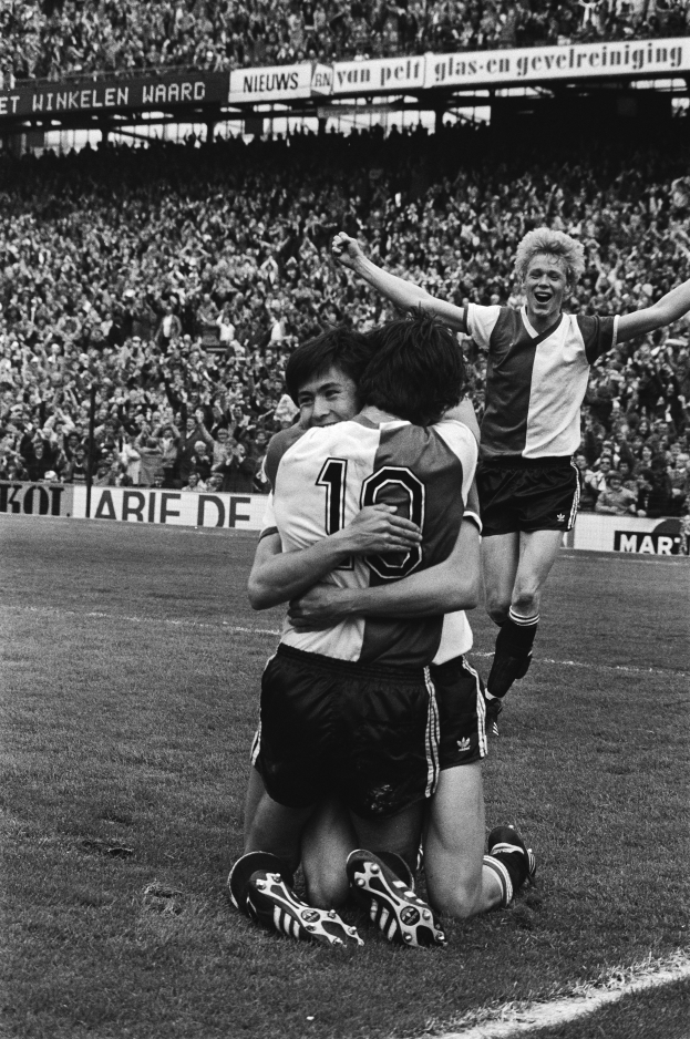 Drei Fußballspieler feiern ein Tor auf dem Boden, umgeben von einer jubelnden Menge in einer schwarz-weißen Stadion-Szene.