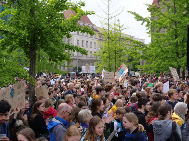 Große Menschenmenge protestiert vor einem Gebäude in Berlin, hält Schilder hoch, mit Bäumen, Fahrzeugen, einem Lautsprecher und Himmel im Hintergrund.