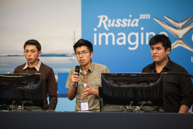 Drei Jungen stehen neben einem Schreibtisch mit zwei Computern, einer spricht in ein Mikrofon, trägt ID-Karten mit roten Markierungen, vor einer Wand mit einer Werbetafel, auf der "Russland Imagine 2013" steht.