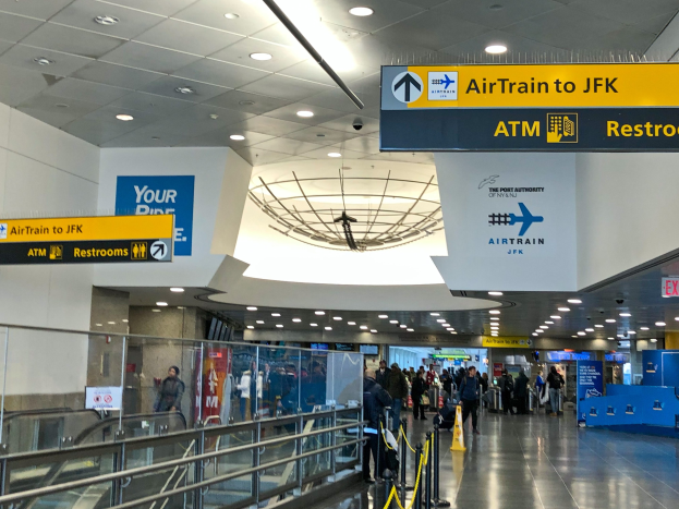 Eine Gruppe von Menschen geht durch eine Flughafenhalle mit einer Rolltreppe auf der linken Seite, Schilder an den Wänden und einem Schild mit der Aufschrift "Airtrain nach JFK".