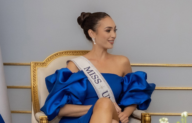 Frau in einem blauen Kleid mit einem "Miss Universe 2019"-Bänder sitzend auf einem Stuhl umgeben von Blumen mit einem Lächeln im Gesicht.