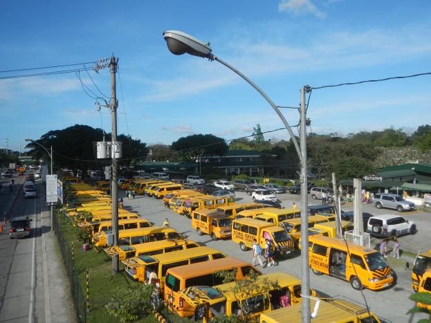 Eine große Anzahl von gelben Schulbussen, die an der Straßenseite geparkt sind, mit Menschen, die auf dem Gehweg gehen, Strommästen mit Drähten, Laternenmästen, Bäumen, Gebäuden und einem bewölkten Himmel im Hintergrund.