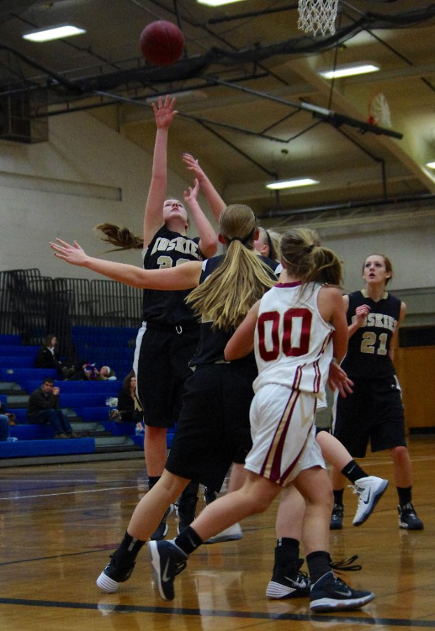 Mehrere Frauen in Trikots auf einem Basketballfeld mit einem Basketball und Netz sowie Zuschauern im Hintergrund an einer Wand mit Deckenbeleuchtung.