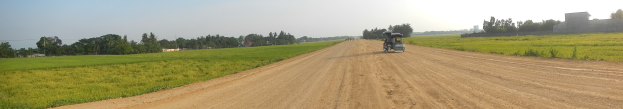 Ein Traktor fährt auf einem Schotterweg durch eine grüne Wiese, umgeben von Bäumen, Gebäuden und einem klaren blauen Himmel.