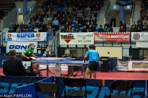 Zwei Männer spielen Tischtennis drinnen mit mehreren Zuschauern, die stehen und sitzen, einigen leeren Stühlen und Werbeplakaten, mit Text in der unteren linken Ecke.
