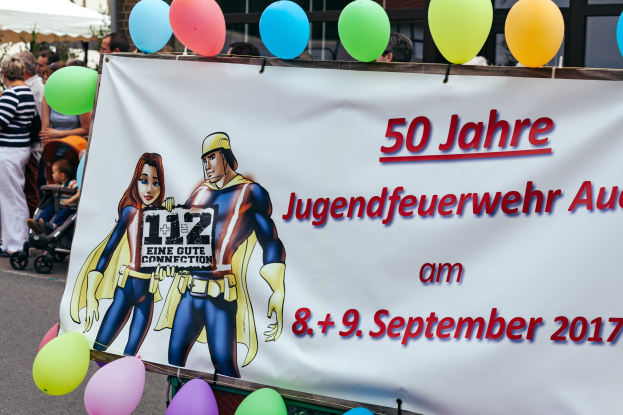 Eine Gruppe von Menschen auf der Straße mit Kinderwagen, ein Banner mit der Aufschrift "50 Jahre Jugendfeuerwehr August 2017" mit Text und Cartoons, ein Zaun, Luftballons, ein Zelt, ein Gebäude mit Fenstern und Bäume im Hintergrund.