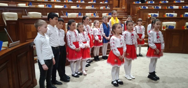 Eine Gruppe von Kindern in weißen und roten Kleidern steht in einem Raum mit einem Podium, Mikrofon, Stühlen, Tischen und Gegenständen im Hintergrund, die Fahnen tragen.
