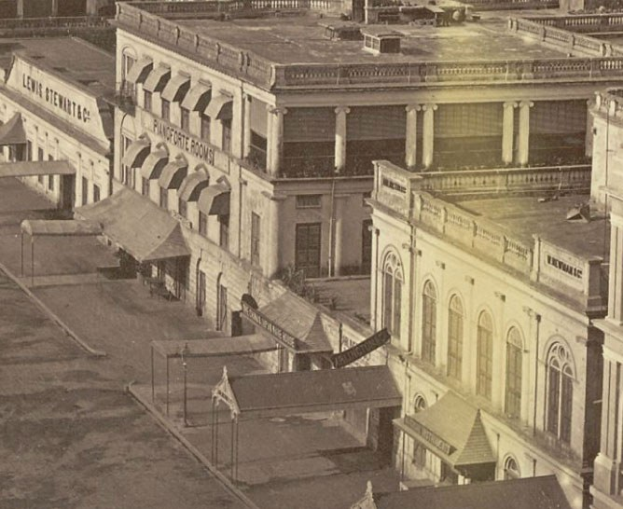 Altes Schwarz-Weiß-Foto einer Stadtstraße mit Gebäuden, die Fenster, Säulen und Namensschilder sowie Schuppen und Pfähle aufweisen.