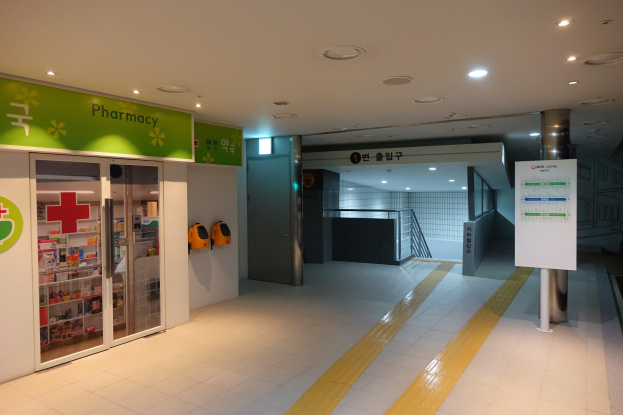 Eingang zu einer Apotheke mit einem "Apotheke"-Schild links, einer Tafel mit Text rechts, beleuchteten Lampen oben und undeutlichen Hintergrundobjekten.