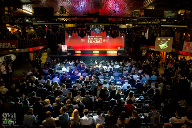 Eine große Menschenmenge sitzt auf Stühlen vor einer Bühne mit Bildschirmen und Bannern, beleuchtet von Scheinwerfern und umgeben von Geländern, gespannt auf den Beginn eines Online-Marketing-Rockstars-Events, mit einem Wasserzeichen in der linken unteren Ecke.