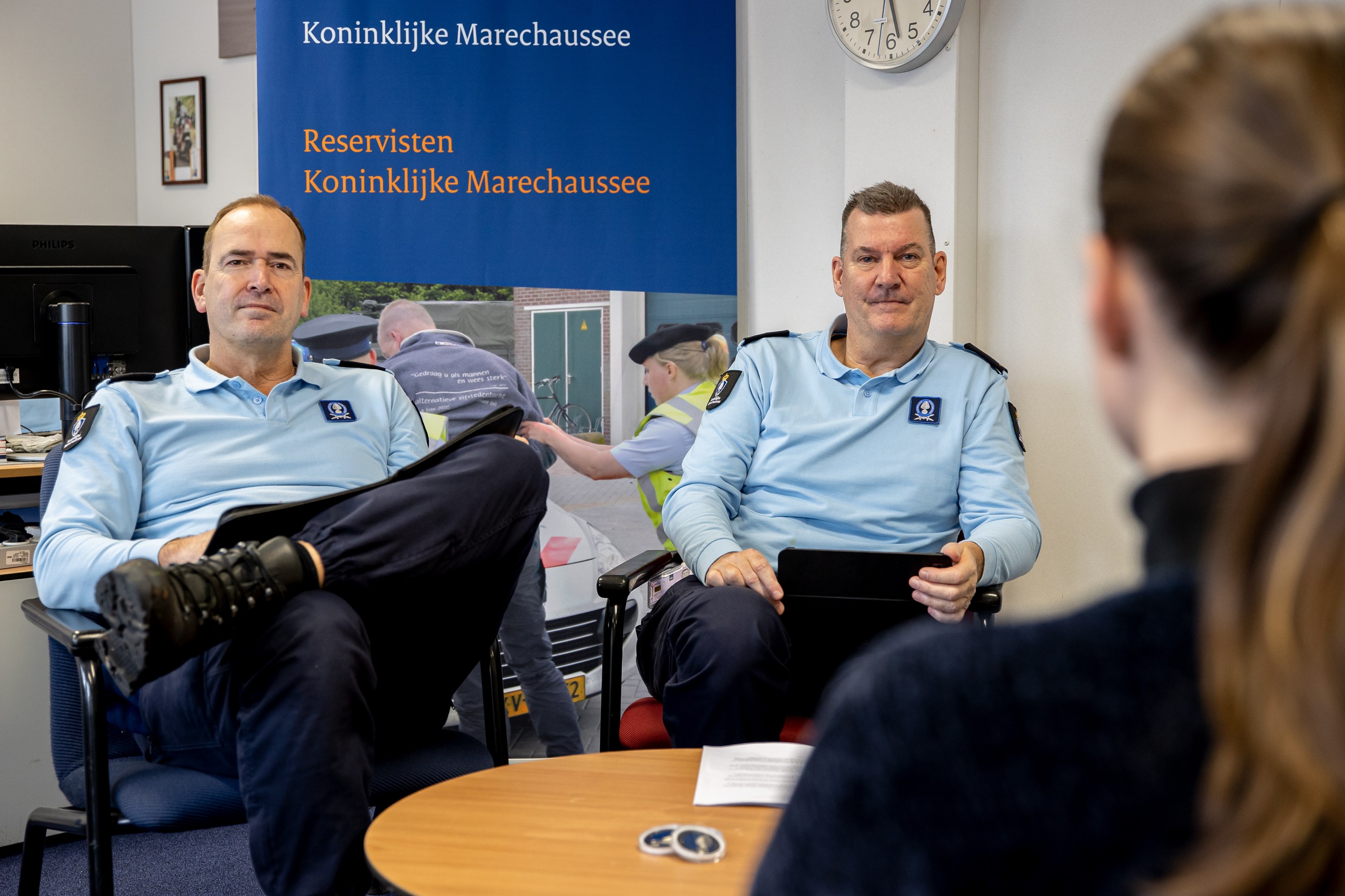 Zwei Polizisten sitzen an einem Tisch mit einer Frau, sie sehen Dokumente durch; im Hintergrund sind eine Wandtafel, eine Uhr und Fotoalben sowie ein zweiter Tisch mit einem Monitor und Zubehör zu sehen.