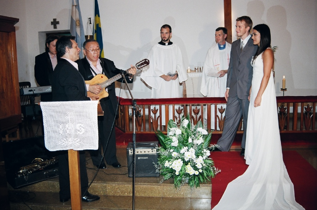 Eine HochzeitsSzene in einem Raum mit zwei Pastoren, einer Braut, einem Bräutigam und drei Musikern; ein Podium mit Stoff vor den Musikern; ein Tisch mit Kerzen hinter den Pastoren; eine Kerze in einem Halter hinter dem Paar; ein Bukett, Lautsprecher, Taschen und roter Teppich auf dem Boden; sowie Fahnen, eine Wand mit einem Kreuz, einen Schrank und einen Stuhl im Hintergrund.