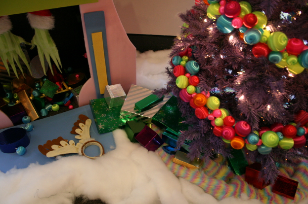 Eine Indoor-Szene mit einer Wand im Hintergrund, einem Baumwoll- und Wolltuch am Boden, einem geschmückten Weihnachtsbaum rechts, Geschenkboxen und dekorativen Gegenständen in der Mitte und einer Hütte mit Dekorationen und Baumwolle oben links.
