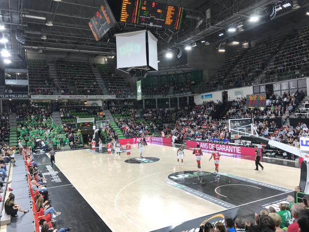 Basketballspiel in einer großen Halle mit Zuschauern, mit einem Spiel zwischen Asvel und Galatasaray, wie auf den Tafeln zu lesen ist.