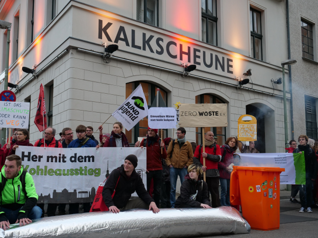 Eine Gruppe von Menschen mit Schildern und Plakaten vor einem Gebäude bei einer Demonstration in Deutschland, mit zwei Personen auf einem Gegenstand sitzend im Vordergrund und einem Mülleimer rechts.