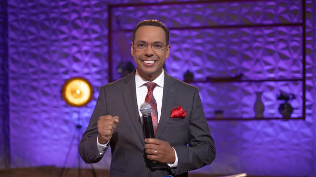 Stephen Colbert, der einen Anzug, eine Krawatte und eine Brille trägt, steht auf einer Bühne und hält ein Mikrofon in der Hand, mit einer Wand, einem Ständer, einem Licht und verschiedenen Gegenständen dahinter.