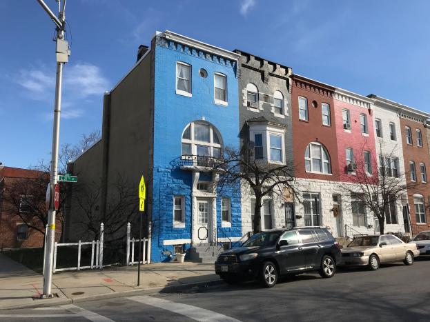 Eine Reihe bunter Häuser säumt eine Straßenkreuzung mit parkenden Autos, Bäumen, Strommasten, einem Zaun und einer bewölkten Himmel, die Baltimores Immobilienmarkt darstellt.