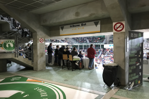 Eine Gruppe von Menschen steht vor einem sitzenden Publikum in einem Stadion während eines Boston Celtics vs New York Jets-Events, mit Schildern, einem Müllcontainer, einer Tafel, einer Treppe und Deckenlampen.