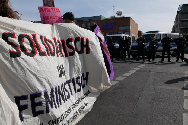 Eine Gruppe von Menschen marschiert auf einer Straße, hält ein Banner mit der Aufschrift "Solidarität und Feminismus", mit parkenden Fahrzeugen am Straßenrand und Gebäuden im Hintergrund unter einem klaren blauen Himmel.