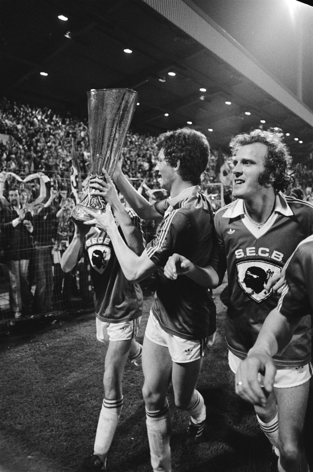 Schwarzes und weißes Foto von Fußballspielern, die mit einem Pokal auf einem Feld feiern, umgeben von einer Menge hinter einem Zaun unter Stadionbeleuchtung.