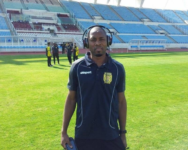 Ein Mann in einem Headset, wahrscheinlich der Trainer der ghanaischen Nationalmannschaft, steht auf einem Fussballfeld in der Nähe eines Stadions und hält ein Objekt, während im Hintergrund einige Menschen zu sehen sind und der Himmel sichtbar ist.