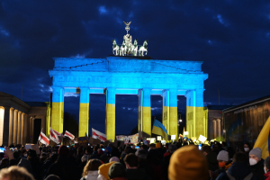 Menschenmenge mit Fahnen und Schildern vor dem Brandenburger Tor in Berlin, mit einer Fahne auf der rechten Seite.
