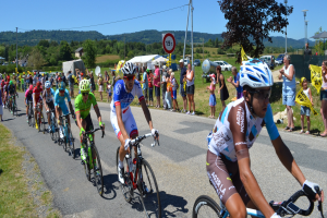 Eine Gruppe von Radfahrern fährt eine Straße entlang, während Zuschauer am Straßenrand stehen, einige tragen Helme und Schutzbrillen oder halten Fahnen, mit Bäumen, Fahrzeugen, Zelten, Gebäuden und Hügeln im Hintergrund unter einem klaren blauen Himmel.