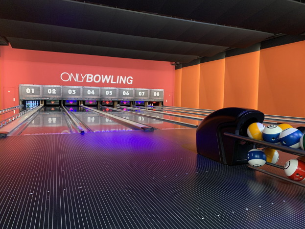Eine Bowlingbahn mit Bowlingkugeln auf der rechten Seite, Kegeln in der Mitte und einer Wand mit Text im Hintergrund, unter einer sichtbaren Decke.