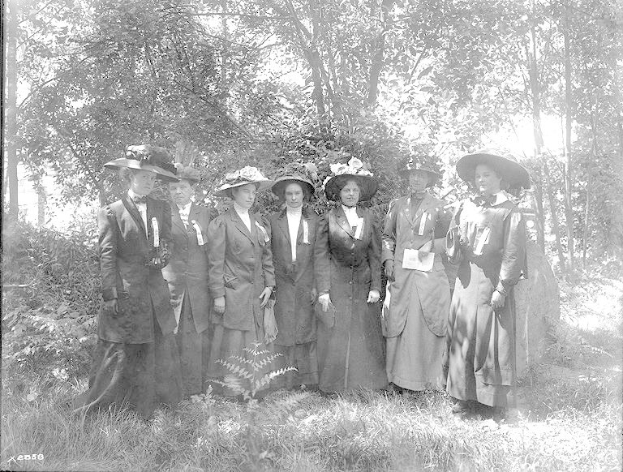 Eine Gruppe von Frauen in Hüten, die zusammen in einem Waldgebiet stehen, als Suffragetten gekleidet, schwarz-weiß.