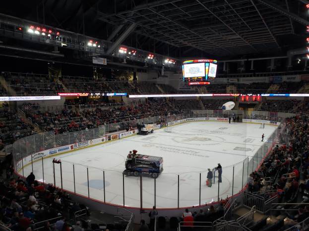 Ein Hockey-Spiel in einer großen Arena mit Fahrzeugen auf dem Eis, Zuschauern rund um die Bahn und Menschen auf den Tribünen, mit Brettern, Texten und Lichtern im Hintergrund.