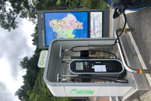 Eine Ladesäule für Elektrofahrzeuge am Straßenrand mit einem Motorrad in der Nähe, ein digitales Anzeigetafel, die anzeigt, dass es eine E-Bike-Ladestation ist, mit Bäumen und einem klaren blauen Himmel im Hintergrund.