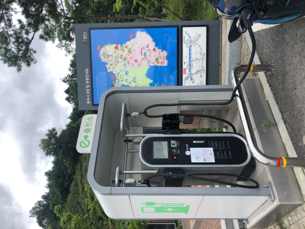 Eine Ladesäule für Elektrofahrzeuge am Straßenrand mit einem Motorrad in der Nähe, ein digitales Anzeigetafel, die anzeigt, dass es eine E-Bike-Ladestation ist, mit Bäumen und einem klaren blauen Himmel im Hintergrund.