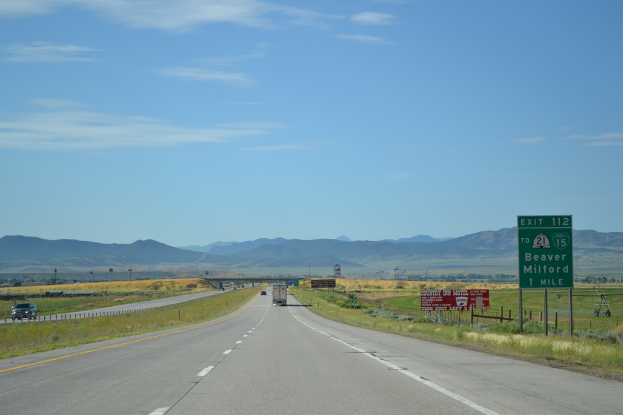 Eine Landschaft mit einem bewölkten Himmel, Hügel, einer Straße mit Kraftfahrzeugen, Barrieren, Schildern, Informationsanzeigen, landwirtschaftlichen Betrieben und Grasflächen.