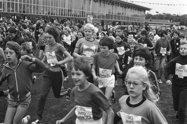 Schwarzes Foto von Kindern, die auf einem Rasenfeld einen Lauf absolvieren, mit einem Gebäude, Bäumen und Pfosten im Hintergrund und einem klaren Himmel.