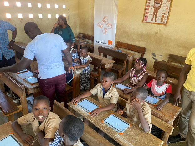 Eine Gruppe von Kindern sitzt an Tischen in einem Klassenzimmer und nutzt Tablets, umgeben von ein paar Menschen, und einem Banner und Plakat im Hintergrund.