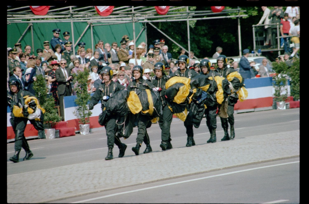 Eine Gruppe von Menschen mit Helmen und Taschen geht während eines Umzugs eine Straße entlang, mit Zuschauern, Pflanzen, Geländern, Pfählen, Fahnen und Bäumen im Hintergrund und Text am unteren Bildrand.