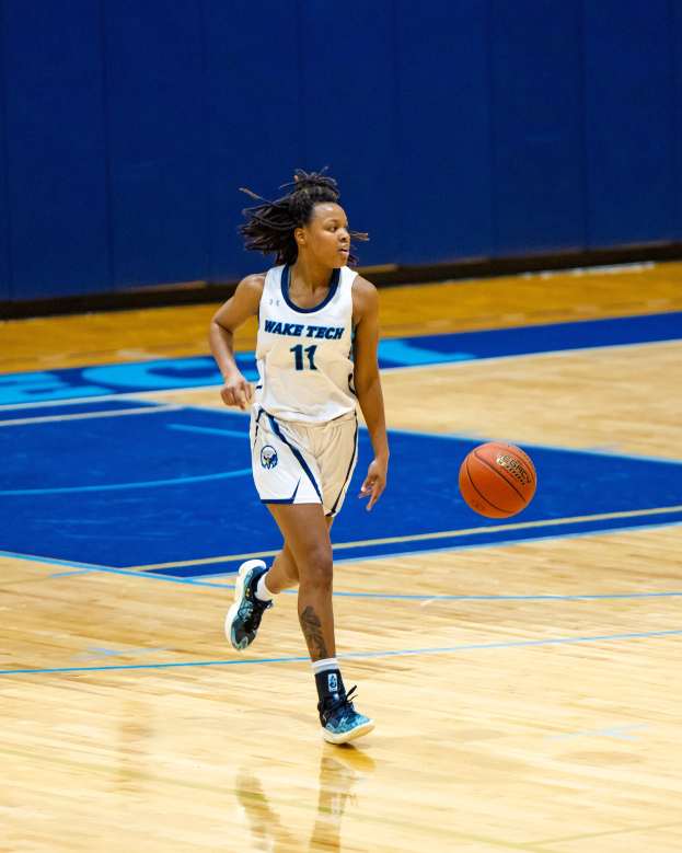 Frau in einem blauen und weißen Basketballtrikot, das einen Basketball auf einem Court dribbelt, trägt ein weißes T-Shirt mit der Aufschrift "Wake Tech Women's Basketball" und blaue Schuhe, mit einer blauen Wand im Hintergrund.