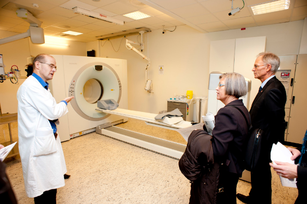 Ein Arzt in einem weißen Kittel erklärt eine CT-Scan-Maschine zwei Menschen in schwarzen Mänteln, die aufmerksam zuhören.