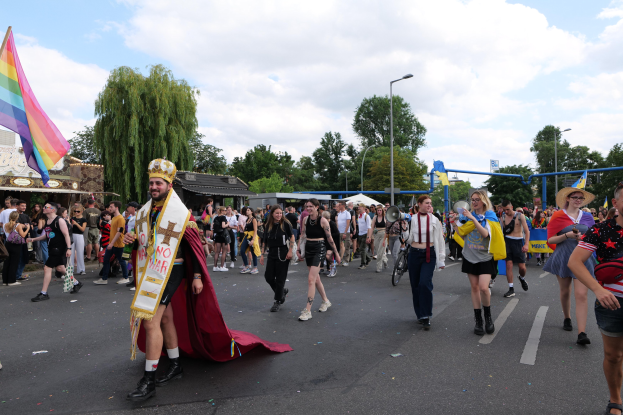Eine Gruppe von Menschen marschiert bei der Pride Parade 2018 mit einer Regenbogenflagge und Musikinstrumenten, im Hintergrund sind Laternenpfähle, Bäume und Hütten zu sehen.